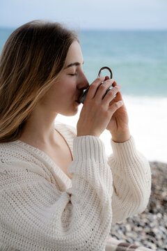 Woman enjoying a warm cup of tea beside the ocean in cool cloudy weather. Cozy slow living, wellness and quiet seaside leisure.