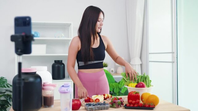 Wellness influencer talking about weight loss diet while filming in modern kitchen. Beautiful Asian woman producing engaging video content about natural healthy eating habits.