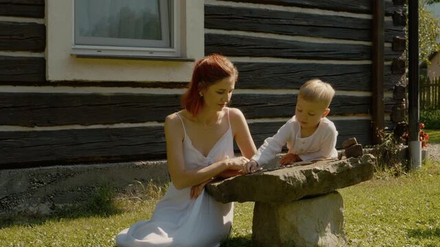 A woman and her little boy spend time in Bucovina. They sit by a traditional house and play with stones on a sunny day, enjoying their time together.