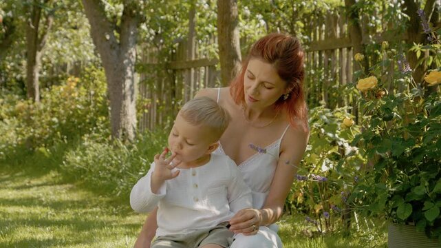 A woman and her little boy have fun in their yard. She puts a raspberry on his fingers and then eats it. They share a moment in the outdoor garden area.