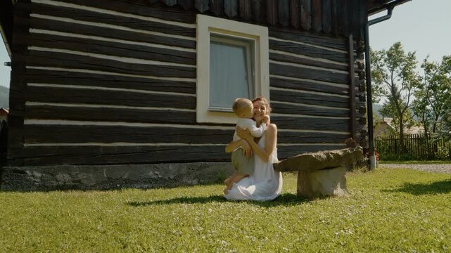 A woman sits on a stone bench while her little boy runs towards her. He hugs her in front of a traditional house in Bucovina during a sunny day.