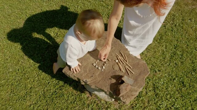 A woman and her little boy enjoy playing with pebbles and crayons outside their house. They arrange the items and create art on a stone.