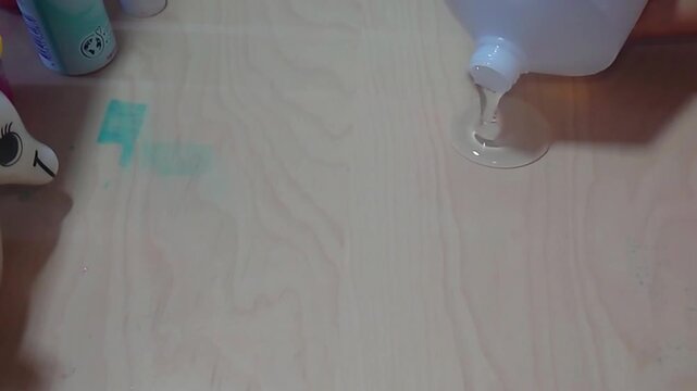 A high-angle overhead shot shows clear PVA liquid glue pouring from a white plastic bottle onto a smooth wooden desk surface to create a base puddle during a fun home STEM science craft experiment.