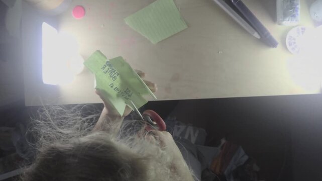 An overhead POV shot shows a preteen child sitting at a bright desk workspace assembling a green paper squishy craft pouch labeled flavored butter using red-handled scissors and crafting supplies.