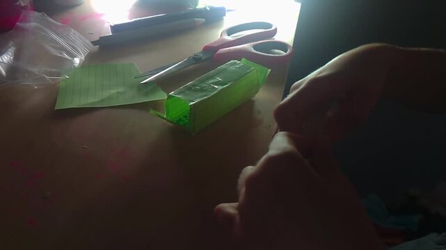A close-up shot shows a preteen child's hands using clear adhesive tape to seal a small rectangular green paper squishy box craft, with scissors and tape rolls nearby on a wooden desktop workspace.