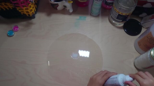 A top-down overhead shot shows a preteen child holding and squeezing a plastic squeeze bottle of liquid activator solution over a clear PVA glue pool on a wooden desk during a home craft science