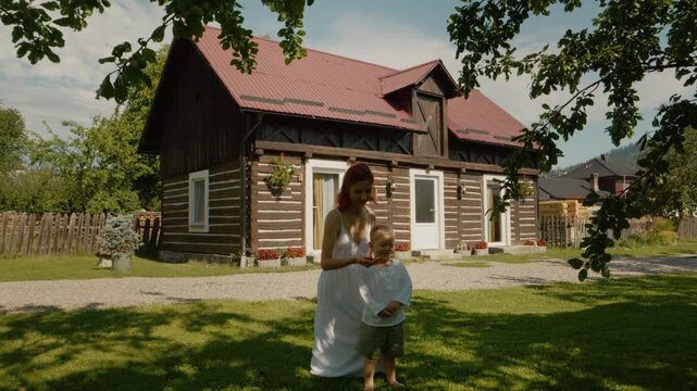 A woman and her little boy are in the yard enjoying time together. They smell flowers and admire nature around their home. It is a sunny day in their backyard.