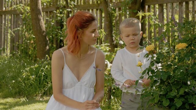 A woman and her young son spend time in their yard. They look at flowers and share smiles as they admire the beauty of nature on a sunny day.