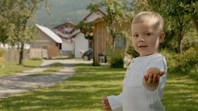 A little boy sits in the backyard holding an apple while enjoying family time. The sun shines on the green grass and houses are seen in the background.