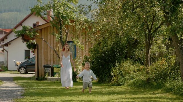 A woman and her young son enjoy their time in the yard. They run and play together while surrounded by trees and a cozy home on a clear day.