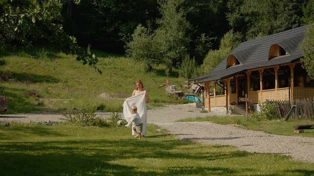 A woman and her little boy spend time in their yard. They play and explore in a green space near their wooden house surrounded by trees.