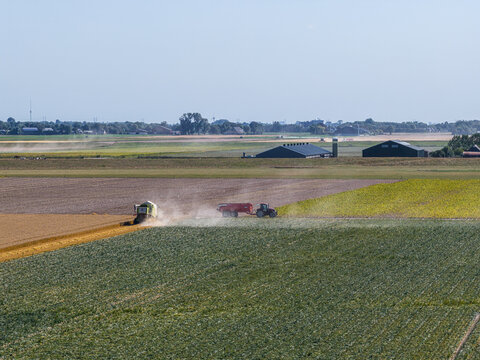 Eemshaven, Groningen, Netherlands - 13 August 2026: Aerial view of a combine harvester and tractor working in fields, creating dust in the flat landscape with farm buildings.