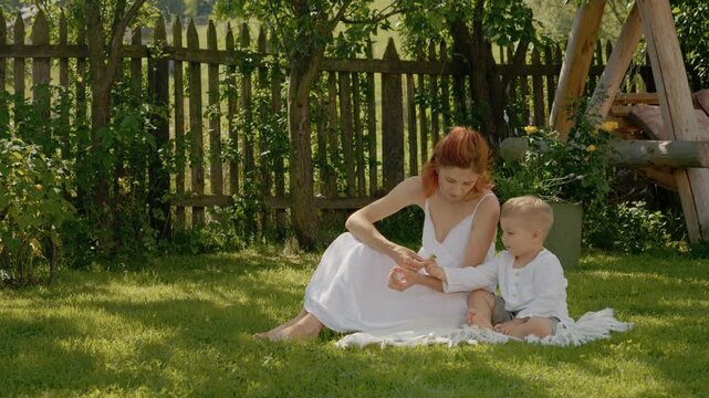 A woman and her little boy sit on the grass in the yard. They enjoy nature as she shows him different flowers. They share moments together in the sunlight.