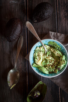 Bowl of crushed avocado, kitchen towel, spoons and whole and hollowed half of avocados on wooden table