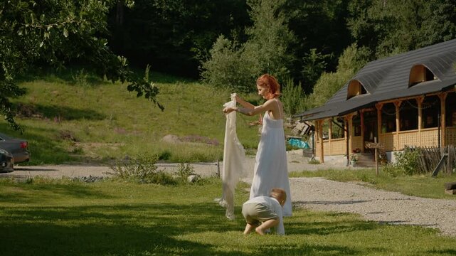 A woman in a dress plays with her little boy outside a traditional house in Bucovina. They have fun together on a sunny day surrounded by nature.