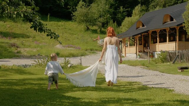 A woman and her little boy walk together in Bucovina. They enjoy their time outside and share moments near a traditional house on a sunny day.