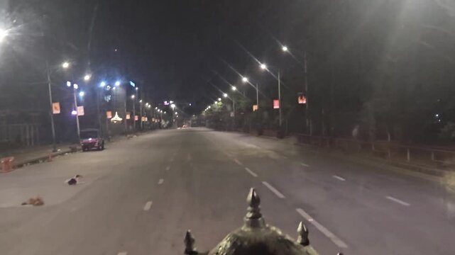 Nighttime driving footage showing a quiet urban road illuminated by streetlights, with trees, buildings, vehicles, and roadside infrastructure. The footage captures a peaceful city street atmosphere