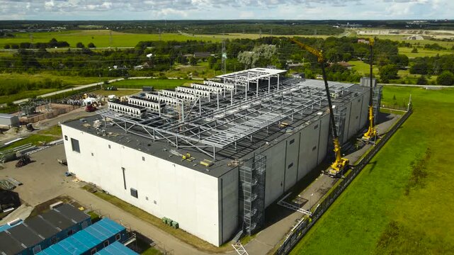 Aerial view showing a modern data center building during its construction phase, with large cranes and heavy machinery operating on the site to install rooftop cooling systems