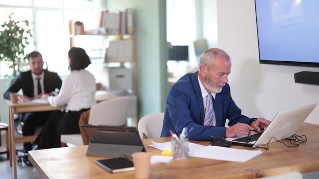 Elderly man sits at desk in office, focused on working with laptop during workday