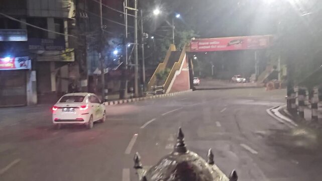 Nighttime driving footage showing a quiet urban road illuminated by streetlights, with trees, buildings, vehicles, and roadside infrastructure. The footage captures a peaceful city street atmosphere