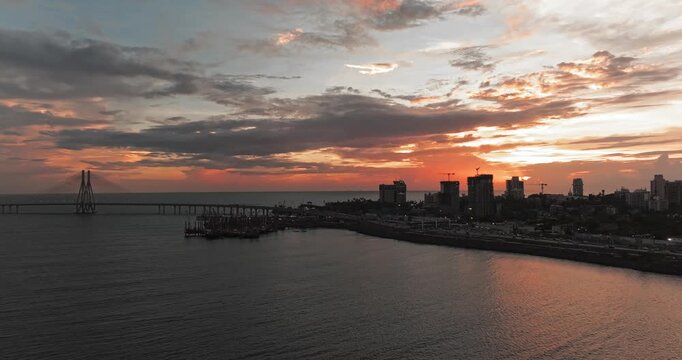 Beautiful sunset view of Mumbai, Maharashtra, with a dramatic orange sky illuminating the city skyline and calm waterfront. The glowing clouds, shimmering water, and striking evening cityscape.