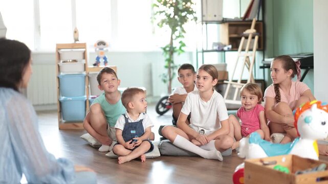 Group of children of different ages sit together on the floor and listen with interest to the teacher against the background of the interior of the kindergarten