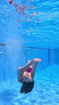 A girl jumps headfirst into the pool and swims mermaid style under the water with a bright, clear blue background, approaches and smiles
