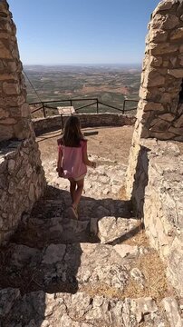 Vertical video following a young girl descending stairs along a dirt and rock path towards a viewpoint high above a large plain full of olive groves with a blue horizon and sky.
