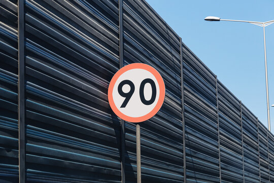 Ninety speed limit sign stands roadside. Dark noise barrier stretches beside roadway. Blue sky surrounds traffic infrastructure. Street lighting rises above wall. Road safety forms background.