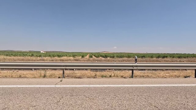 A lateral view of a highway with a metal guardrail, surrounded by fields of green crops and dry grass under a clear blue sky. The video captures the essence of rural road travel in Spain
