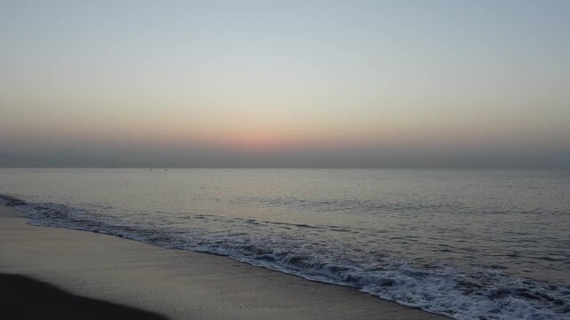 Seashore of sandy beach with waves coming in on a very calm Mediterranean Sea, with clear sky and horizon line, at dawn or dusk
