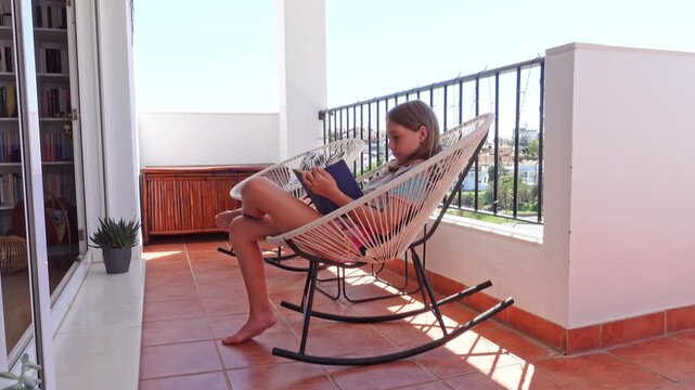 Preteen girl reading a book sitting and relaxing in a modern rocking chair on a terrace on a sunny summer day
