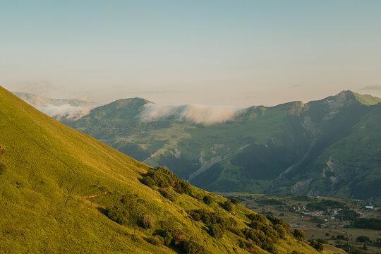 Scenic view of a mountain valley and village in Gudauri, Georgia, with dramatic creeping clouds rolling over a green ridge at golden hour