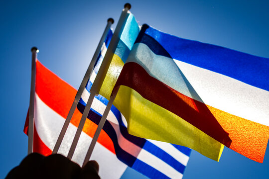 Waving flags stretch toward the sun as brilliant colors dance in the wind. The handheld scene captures teamwork celebration and international connection under a perfect blue canopy