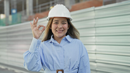 Woman in white hardhat makes ok sign at building site with corrugated metal hoarding and scaffolding, smiling  confidence progress. © Krakenimages.com