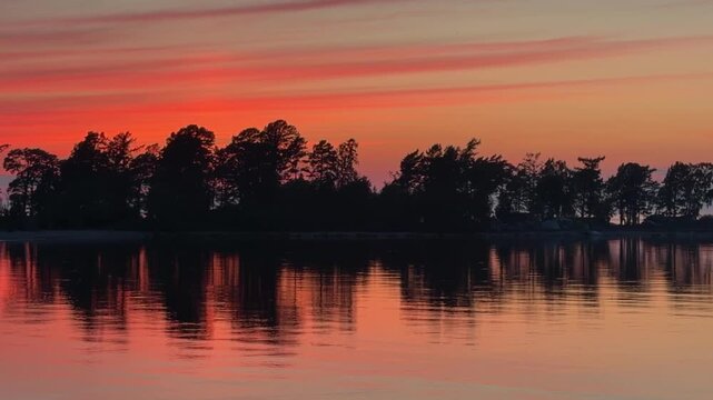 Sunset over calm lake reflecting vibrant orange and pink hues with silhouettes of trees along the shoreline creating a serene natural landscape