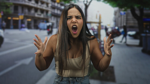 Young woman with clenched fists and a scowling face yelling with raised shoulders on a crowded street, tanktop and long hair visible; anger confrontation.