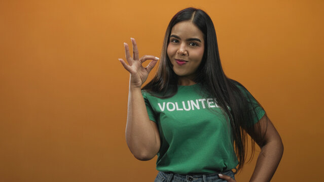 Young hispanic woman makes ok sign with hand in studio wearing green volunteer tshirt against orange backdrop, hand on hip; playful confidence.