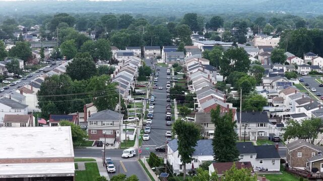 Cars travel down long straight street lined with gabled rooftops in Union, New Jersey, dense rows of houses and parked vehicles stretch beneath tall shady summer trees. Aerial drone wide shot