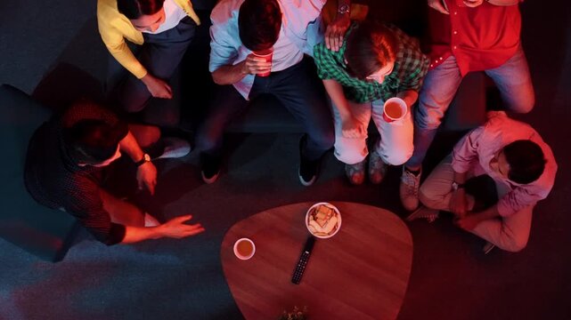 Diverse friend cheering football together while happy man holding snack bowl and remote at dark living room. Caucasian people watching movie and spending time together while eating snack. Convocation.