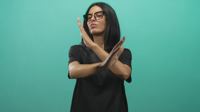 Woman hands crossed in x stop gesture, wearing glasses and black t shirt against turquoise studio backdrop; defiance.