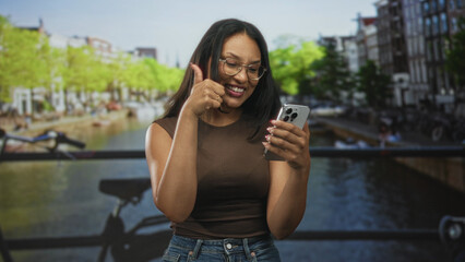 Fototapeta premium Woman tapping smartphone and giving thumbs up with hands visible on street canal bridge wearing glasses and jeans; digital connection happiness.