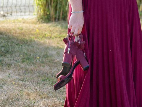 Femme en robe bordeaux tenant ses chaussures &agrave; talons &agrave; la main lors d&rsquo;un mariage en ext&eacute;rieur