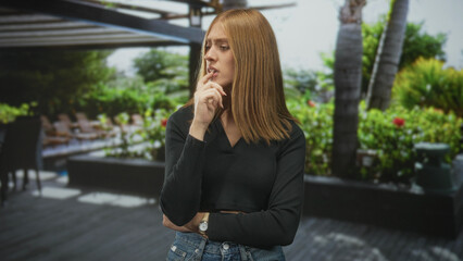 Fototapeta premium Woman with hand on chin touching her jaw, wearing jeans and a watch, standing on a poolside patio near plants and palm trees; introspection concern.