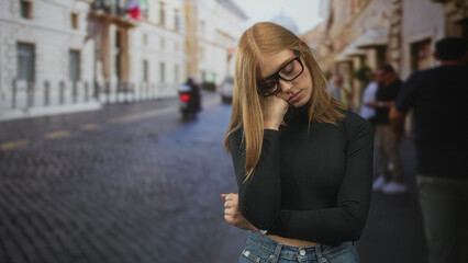 Fototapeta premium Redhead woman with glasses resting chin on fist, eyes closed, standing on a cobblestone street lined with buildings; weariness solitude.