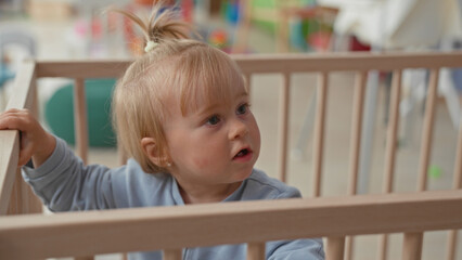 Fototapeta premium Toddler girl with blonde ponytail grips wooden crib rail with small hands, looking sideways near play area in building; quiet curiosity.