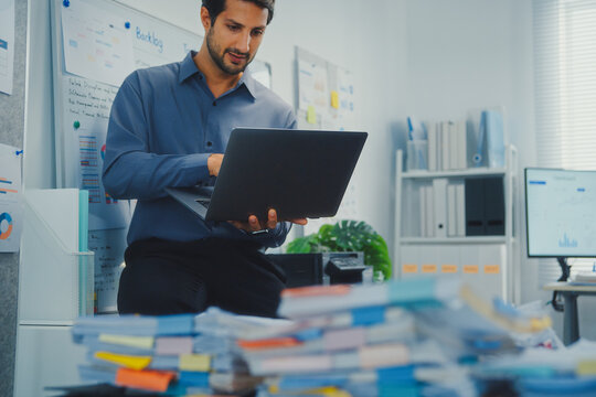 Thoughtful young Latin businessman stand with laptop analyzing data above paperwork stack. Hybrid work audit, digital transformation strategy, business corporate finance, executive decision.