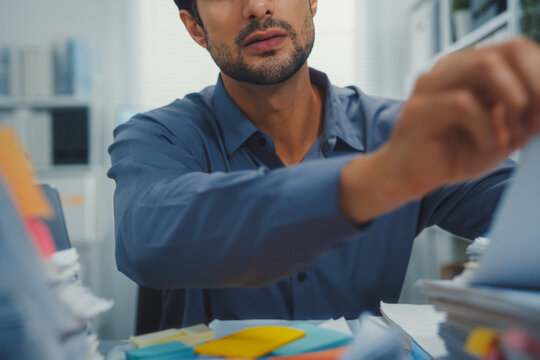 Closeup overworked young Latin businessman reading document in large stack of paperwork file at office desk. Tax audit deadline, accounting stress, corporate burnout, bookkeeping, bureaucracy concept.