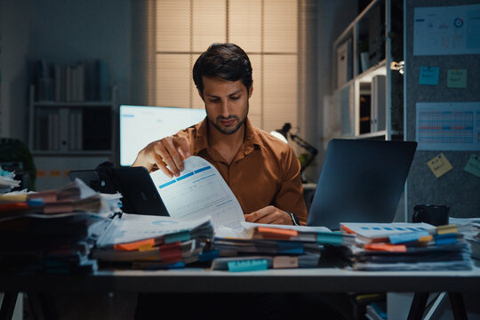 Overworked young Latin businessman working late on document in large stack of paperwork files at office night. Tax audit deadline, accounting stress, corporate burnout, urgent business concept.