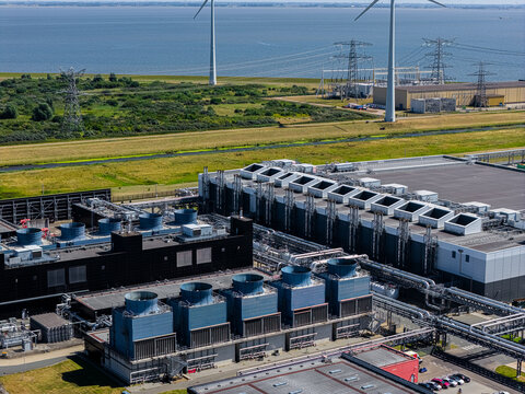 Eemshaven, Netherlands - 13 August 2026: Aerial view of the Eemshaven industrial area featuring large cooling towers, complex piping systems, and wind turbines in the background near the coast.